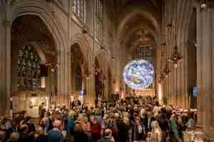 A large crowd of people stood in a church with a floating model of the Earth hanging from the roof in the background
