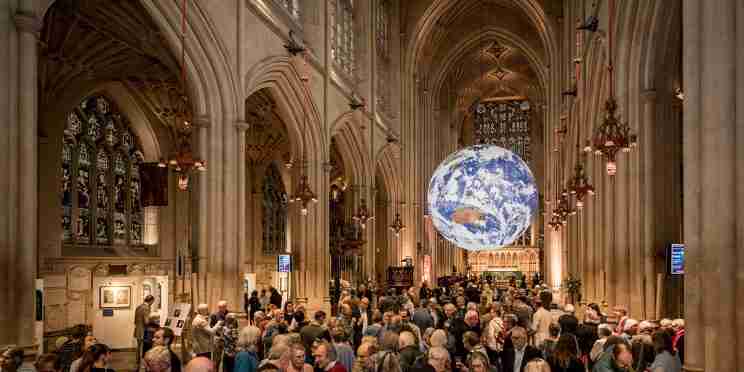 A large crowd of people stood in a church with a floating model of the Earth hanging from the roof in the background