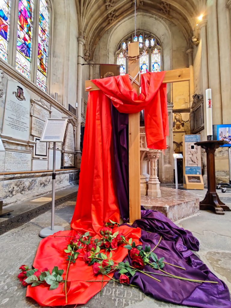 Wooden cross with red and purple fabric draped over, with roses laid on the fabric.